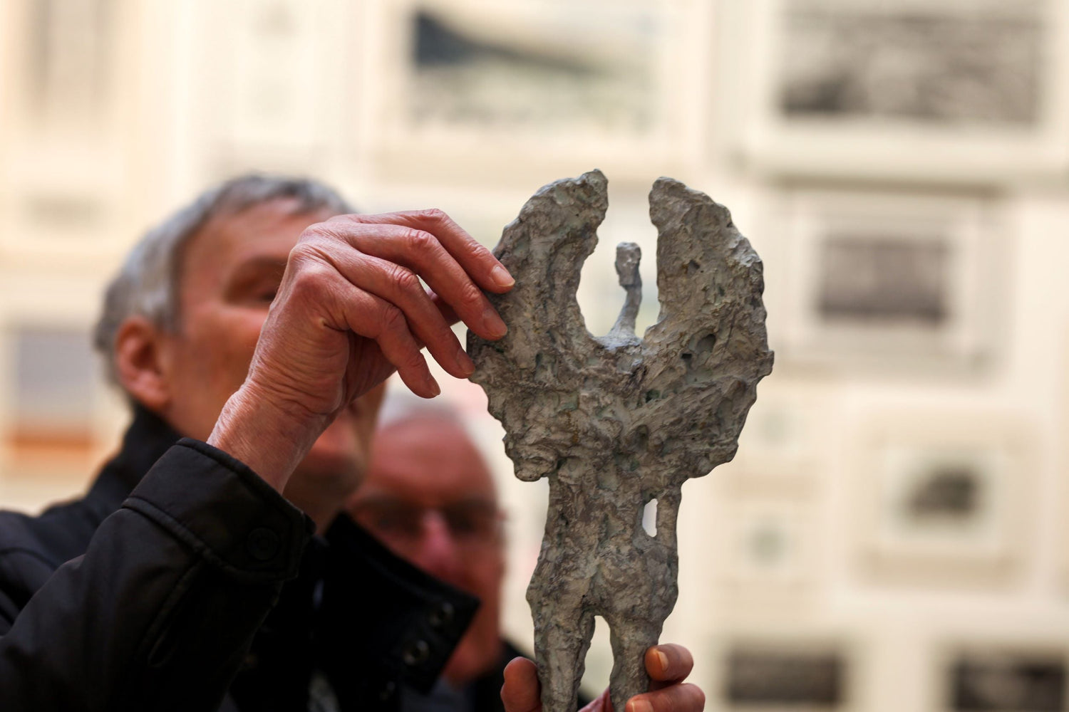 In the foreground, in the centre third of the image, there is an abstract sculpture of a figure with wings. To the left a man with grey hair and black jacket gently touches the sculpture with his thumb and index and middle finger. The background is blurred but we can see the vague outline of artworks hung on a wall.