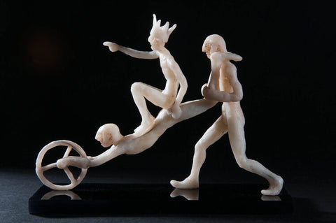 Three pale wax sculptures of people. One wears a crown and points forward, one holds a wheel and one pushes the character holding the wheel. The sculptures are set against a dark background.