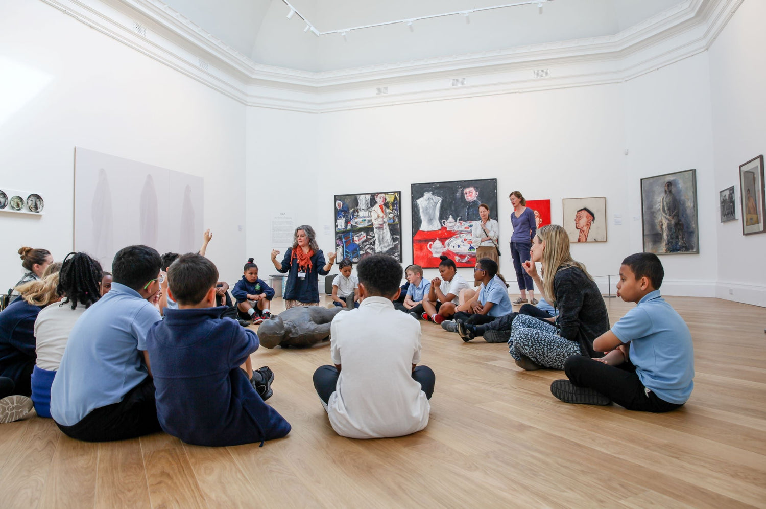 A group of school children sit in a circle surrounding a metal sculpture of a man lying down. They are in a gallery with several artworks hung on white walls. A lady kneels up within the group and gestures to the children. Two people stand behind watching.