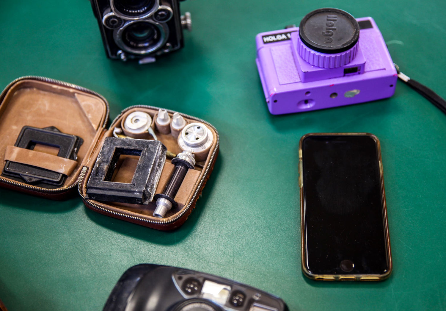 On a dark green surface sit five different types of camera. In the top right corner is a bright purple HOLGA camera with a smartphone below it. At the top and bottom left are two different black film cameras, and between them is an open case with parts for a film camera inside. 
