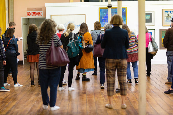 
      A group of people stand facing forward in a gallery space viewing artworks 