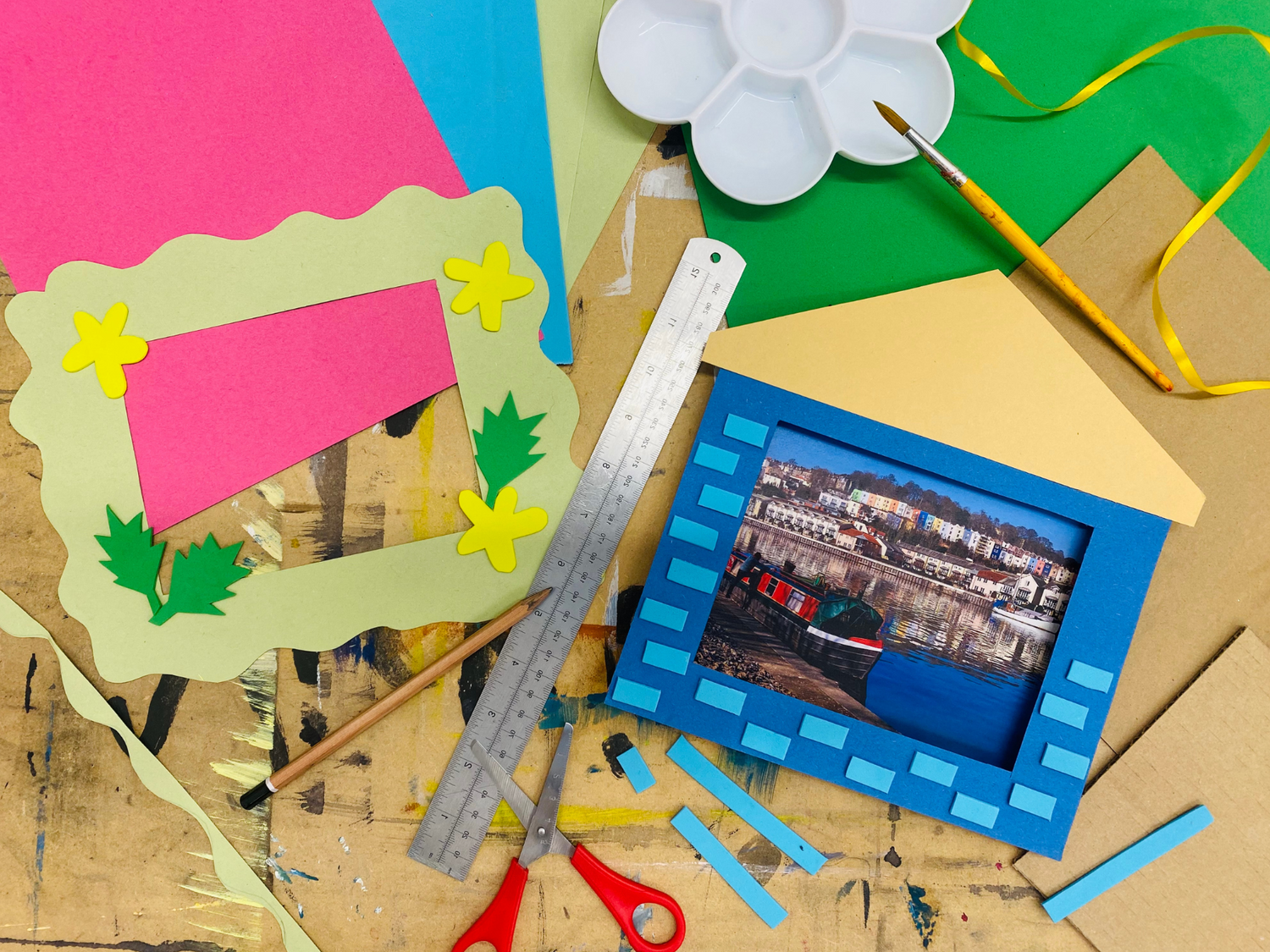 A craft table scattered with materials to make picture frames. There are plies of different coloured card across the top of the image, a paint palette, paint brush and some yellow ribbon in the top right corner, and some scissors, a pencil and a metal ruler in the centre. 
